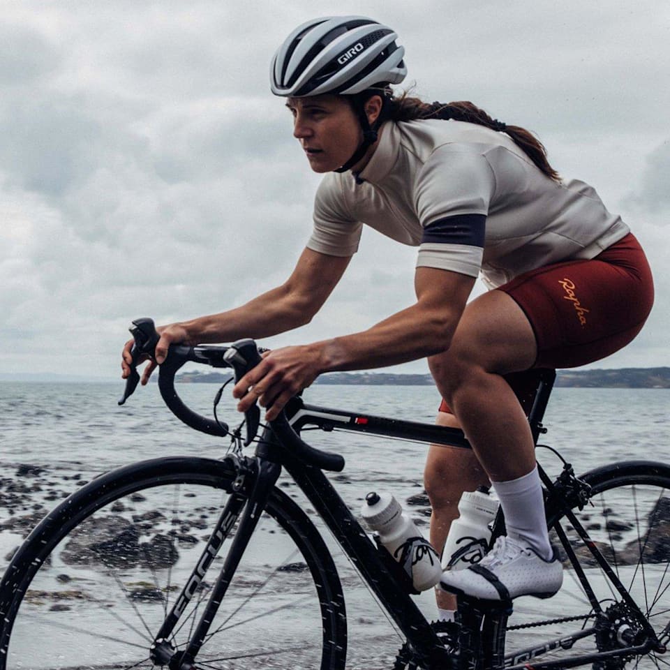 A cyclist clad in Rapha gear intently navigates a coastal road, her ride emphasized by the misty ocean backdrop @ Bici.