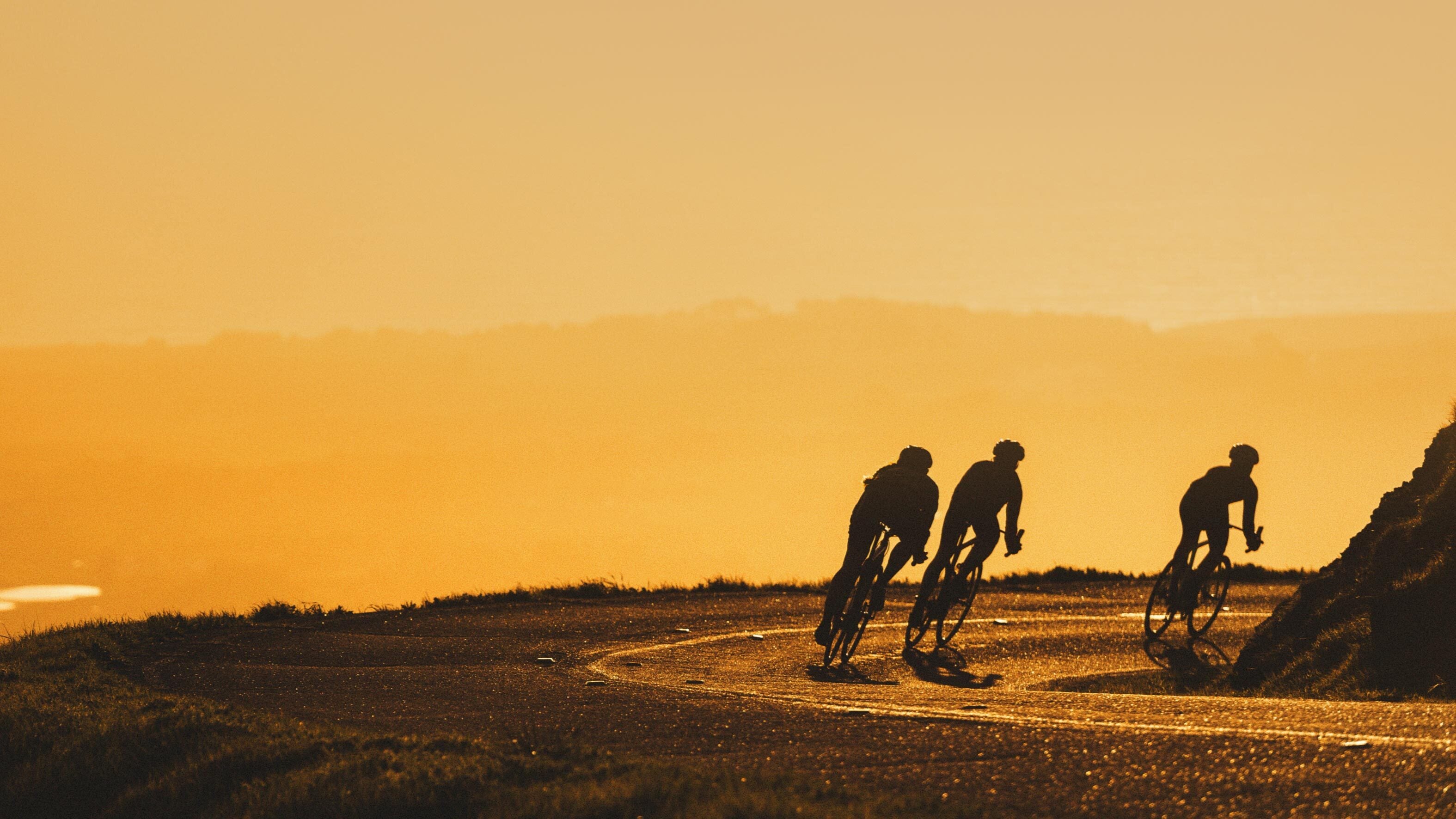 Silhouetted cyclists climbing a hill against an atmospheric, yellow-orange sunrise, evoking the energy of early morning rides @ Bici.