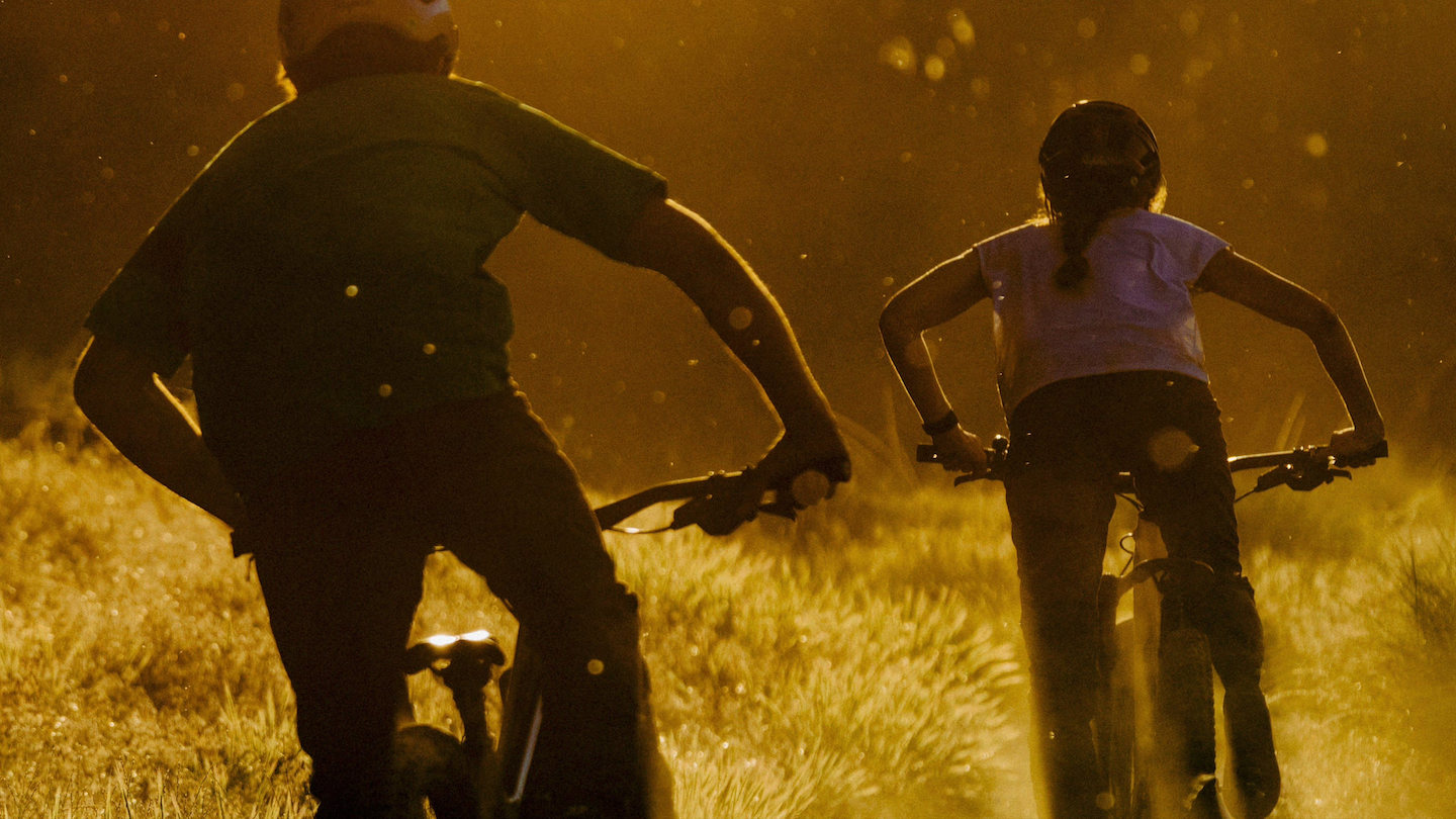 Mountain bikers in a golden-hour glow, riding through a dust-filled trail, capturing the thrill of the ride @ Bici.