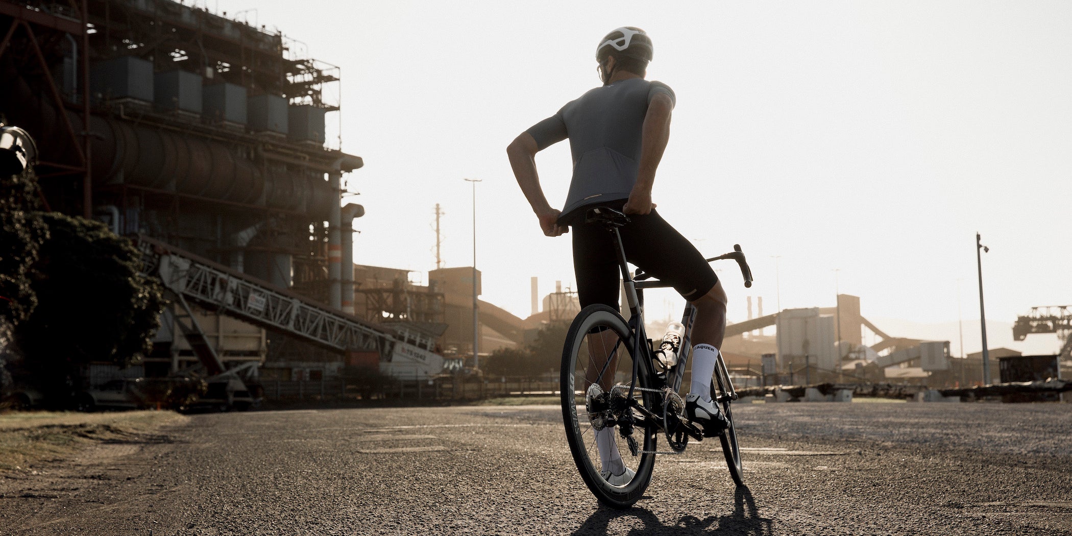 CYCLIST STANDING IN FRONT OF A GRAIN ELEVATOR WITH A BIKE ADJUSTING HIS JERSEY