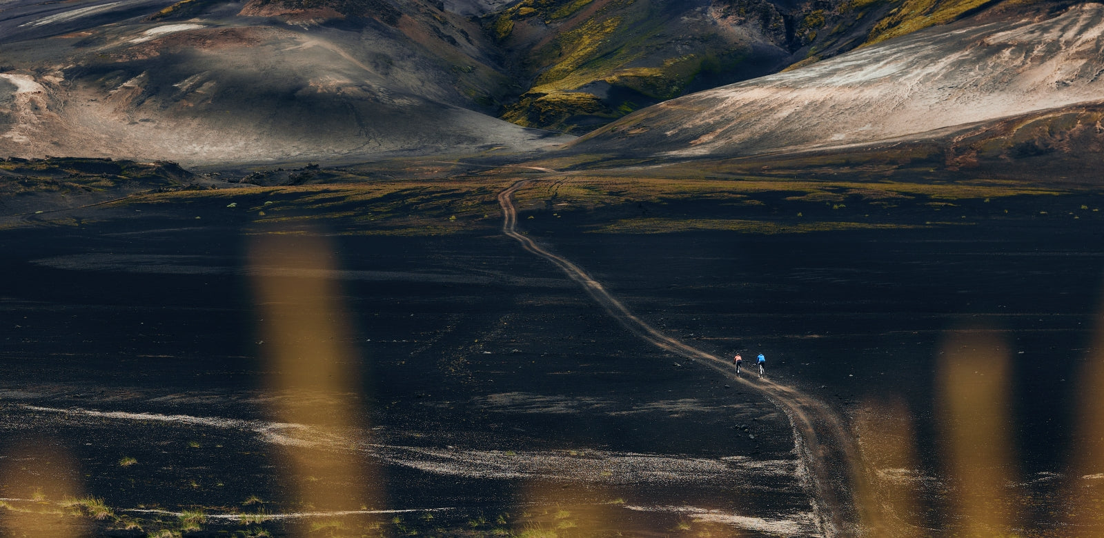 Two riders navigating a winding gravel path through a dramatic, sunlit mountainous landscape @ Bici.