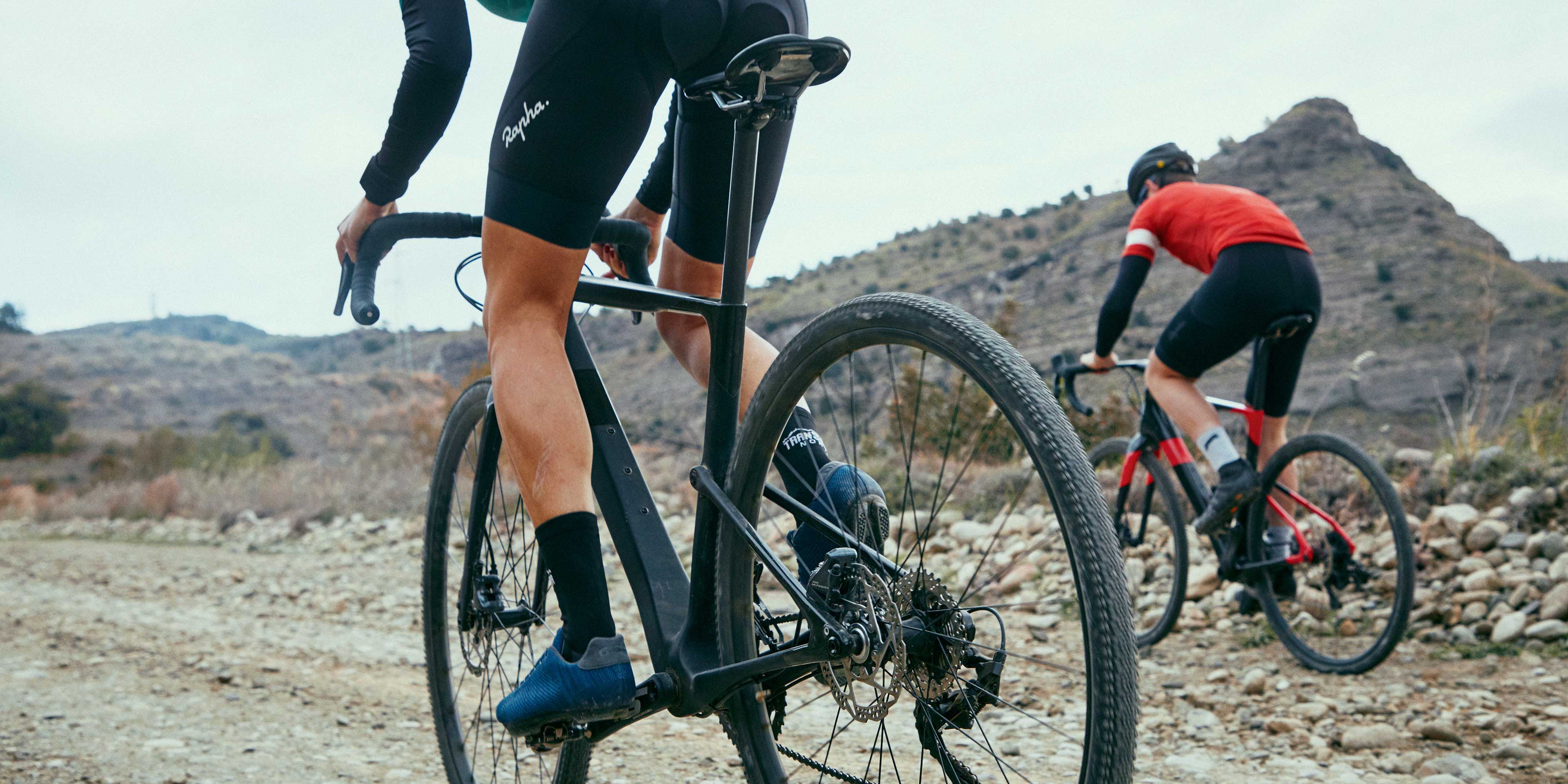 Cyclist in black attire riding a gravel bike on a rocky desert trail, followed by another in the background @ Bici.