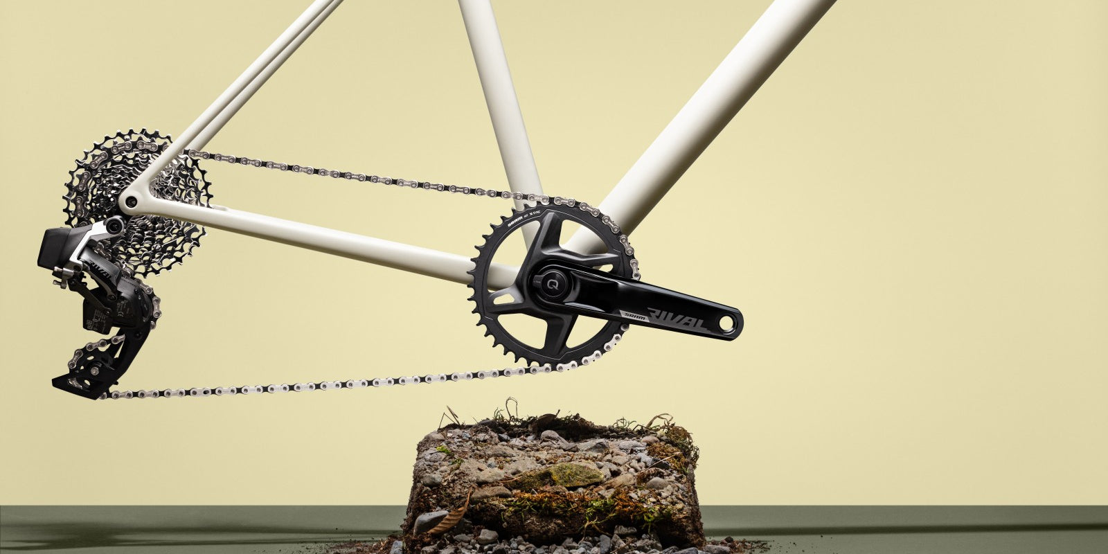Detailed view of a bicycle's crankset and chain, mounted on a white bike frame against a pale yellow backdrop @ Bici.