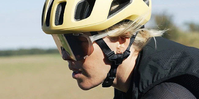 Side profile of a cyclist wearing stylish yellow cycling glasses and helmet, mid-ride against a soft-focus natural backdrop @ Bici.